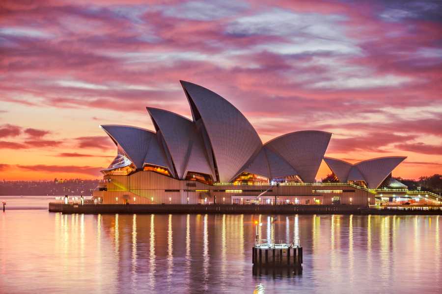 The Sydney Opera House at dusk as the pink clouds, peach sky and golden lights reflect in the water.
