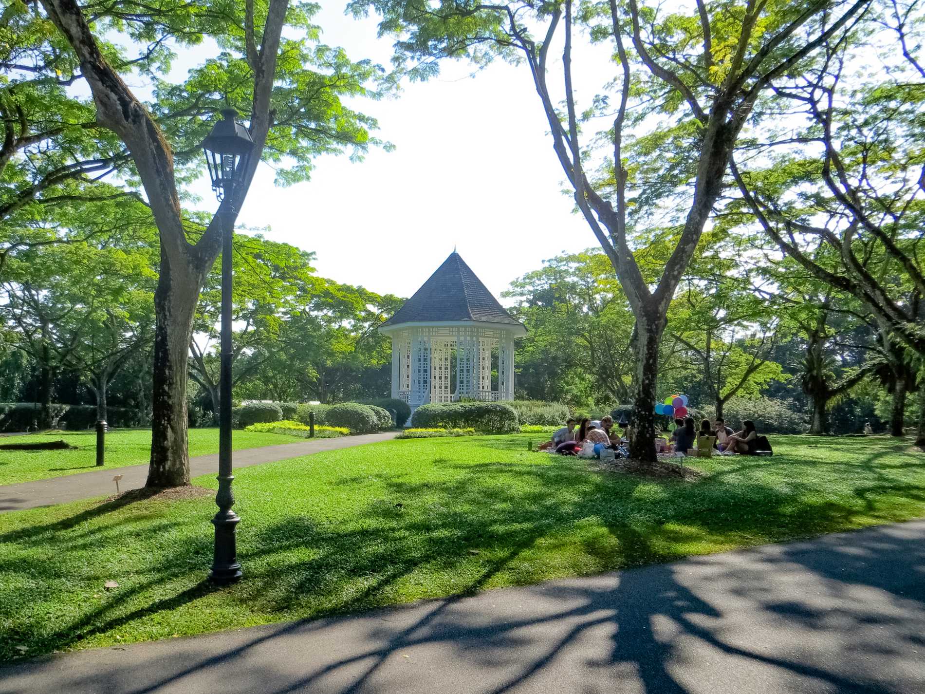A group of friends celebrate with a picnic and balloons in front of the white bandstand in the Singapore Botanic Gardens. 