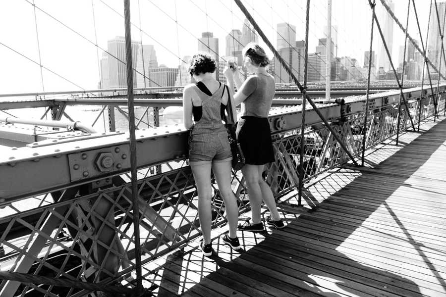 Two girls taking photos of NYC Harbor from the boardwalk on Brooklyn Bridge with the skyscrapers of NYC in the background.